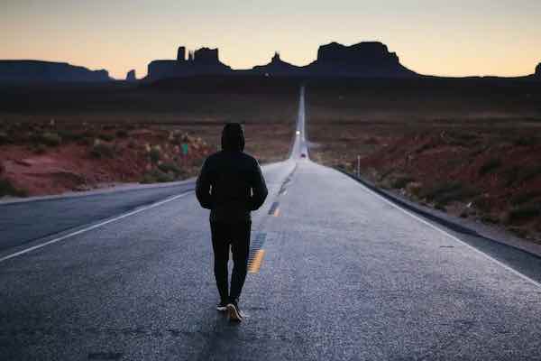 A silhouetted person walking alone down a long, straight highway leading toward Monument Valley's distinctive rock formations at sunset/dusk.