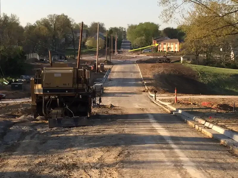 Sunlit road under construction, bordered by trees and utility poles, with machinery in the foreground. Buildings and greenery are visible in the distance. In the distance my apartment quadraplex is shown with a yellow arrow pointing to the window of my of