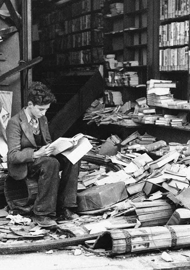 A young man sits amidst a pile of disordered books outside a damaged bookstore, deeply engaged in reading. The scene captures a sense of calm amidst chaos.