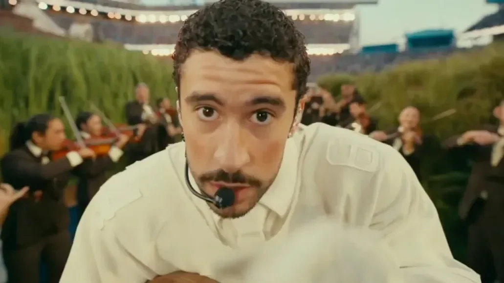 Performer Bad Bunny wearing a headset and white shirt stands close to the camera, with an orchestra playing in the blurred background, creating an energetic atmosphere.