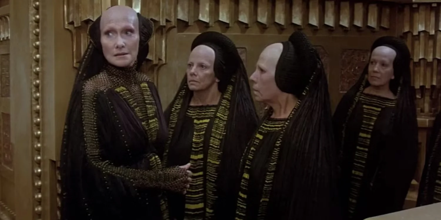 Four Bene Gesserit priestess's stand in ornately detailed robes with dark veils, their expressions serious. The room's golden tones add an austere, ceremonial ambiance.