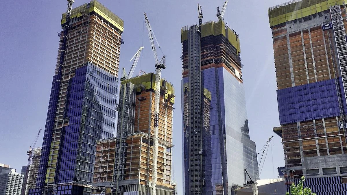 Four skyscrapers under construction with cranes against a clear blue sky. The buildings have glass exteriors, steel frames, and an urban setting.