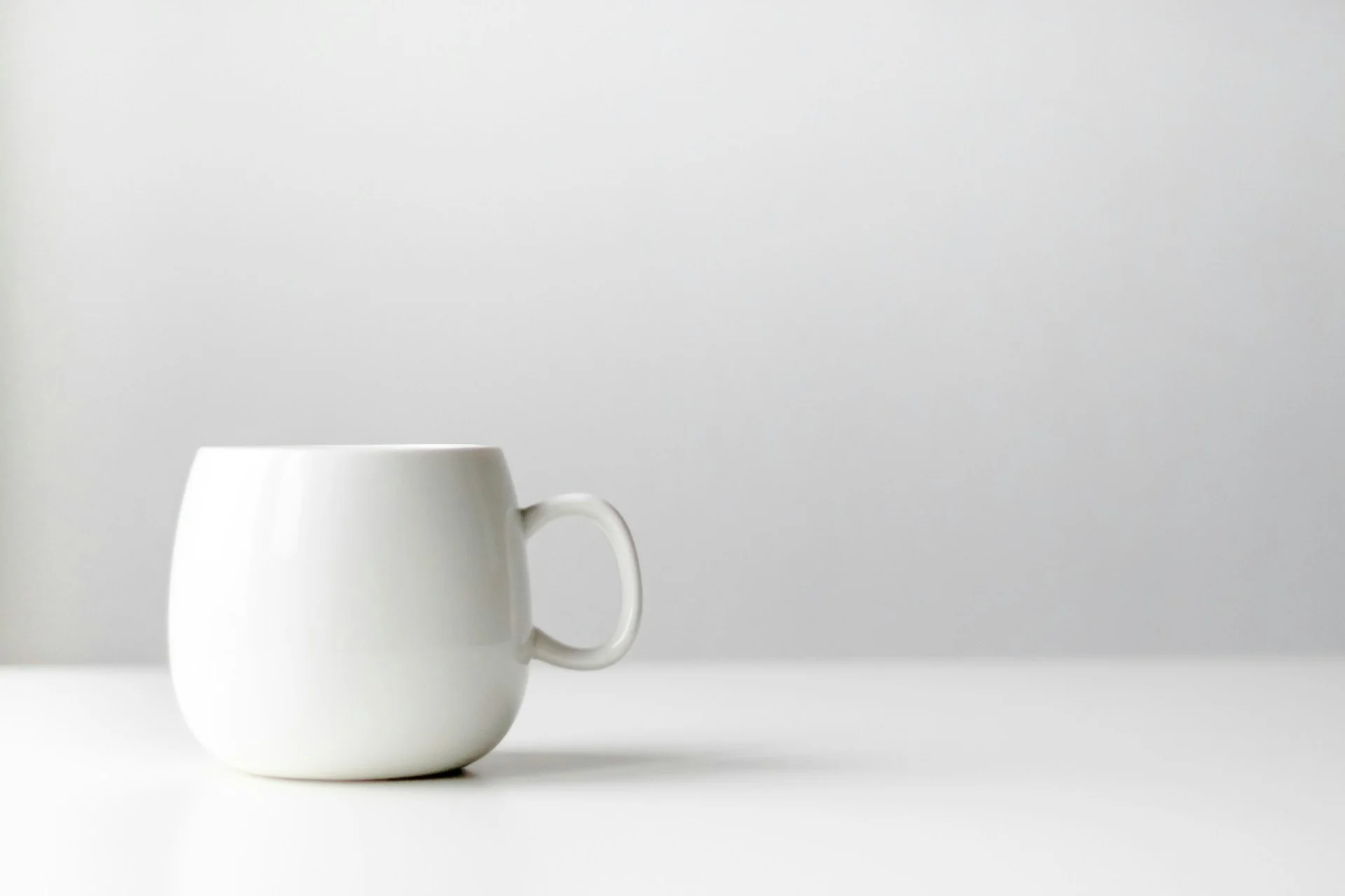 A plain white mug with a rounded shape sits on a minimalist white surface against a soft gray background, evoking a calm and simple atmosphere.