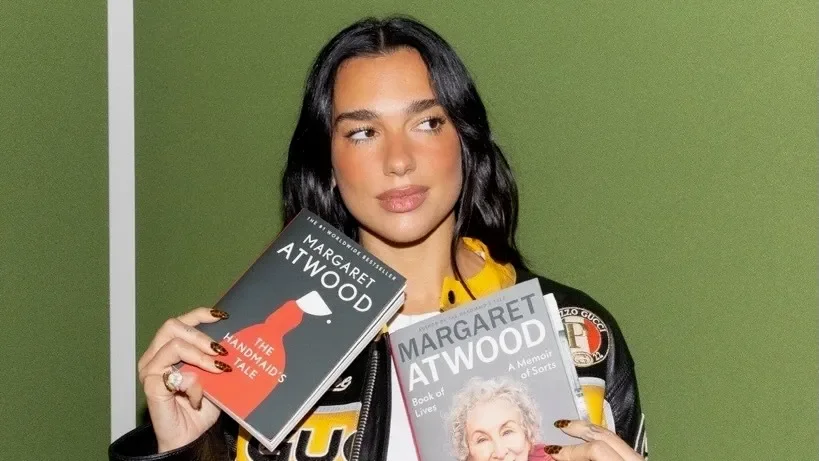Dua Lipa holds two books by Margaret Atwood, 'The Handmaid's Tale' and 'Book of Lives', against a green background.