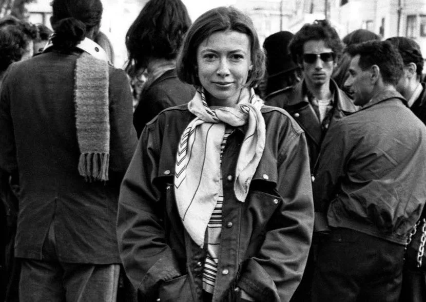 Black and white photo of a younger Joan Didion. She is facing the camera on a crowed sidewalk.