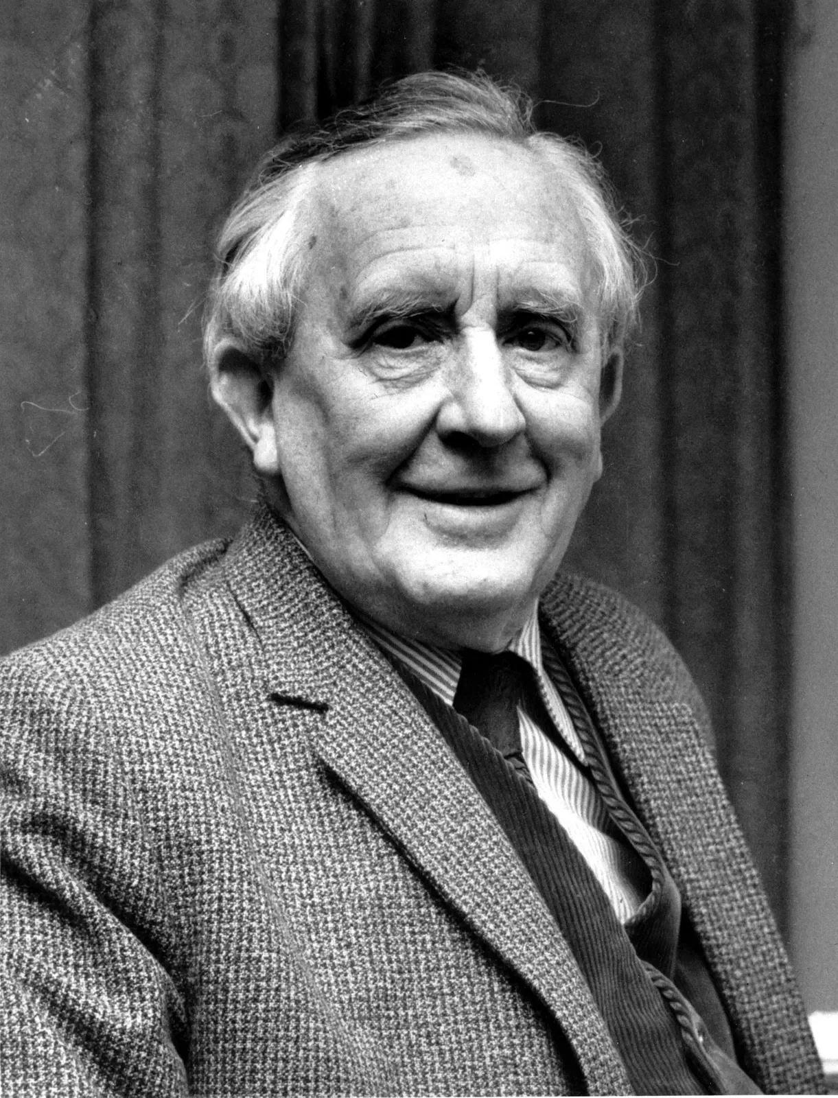 A smiling J.R.R. Tolkien in a tweed jacket and tie sits indoors. His expression is warm and relaxed, conveying a sense of wisdom and approachability.