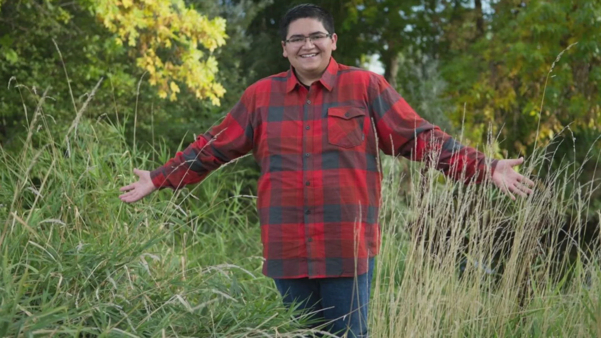 Kendrick Castillo wearing a red and black checkered shirt stands in a field with tall green grass, smiling and spreading their arms wide, conveying joy.