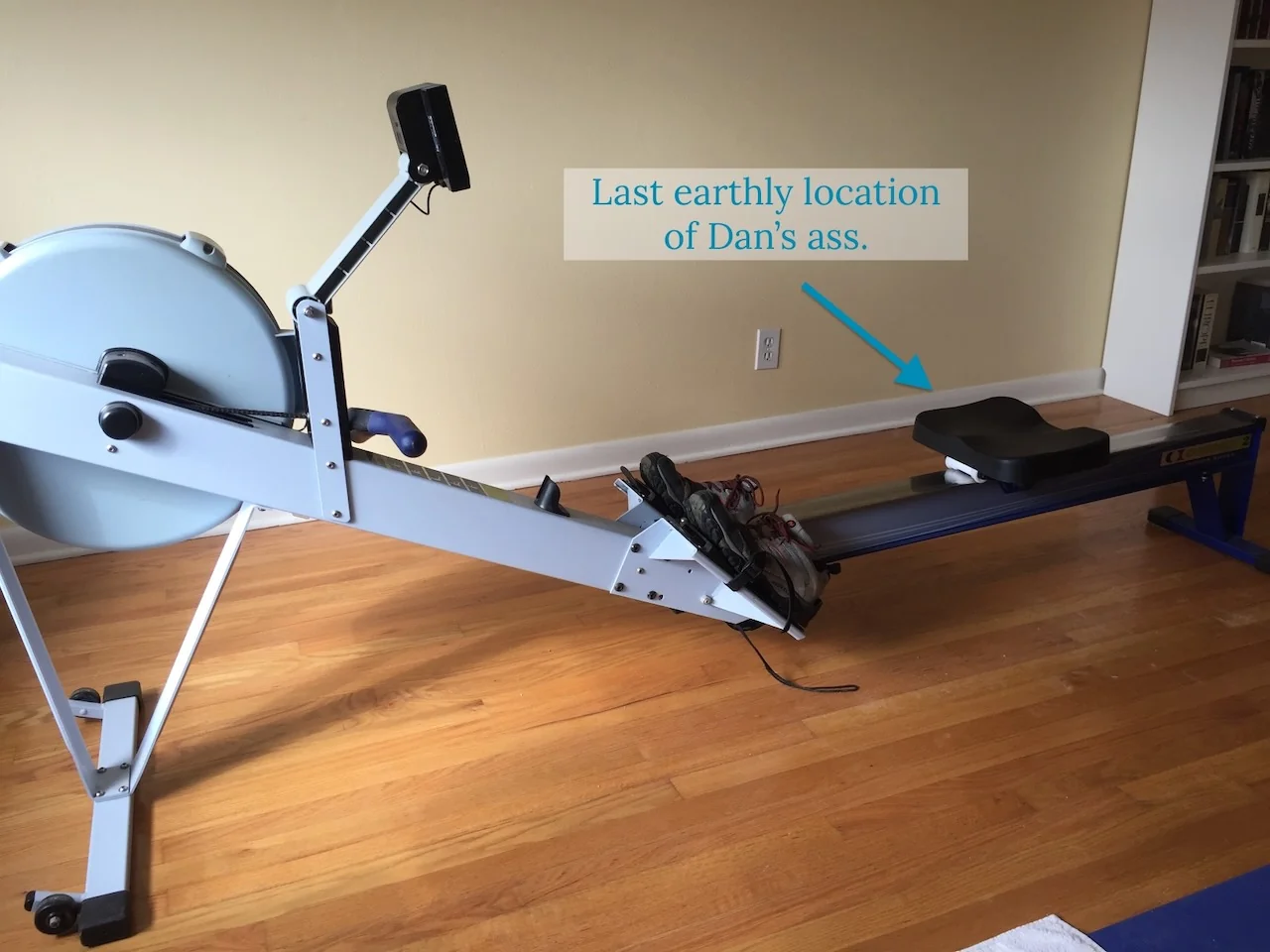 Indoor rowing machine on wooden floor with humorous text overlay pointing to the seat, reading "Last earthly location of Dan’s ass."