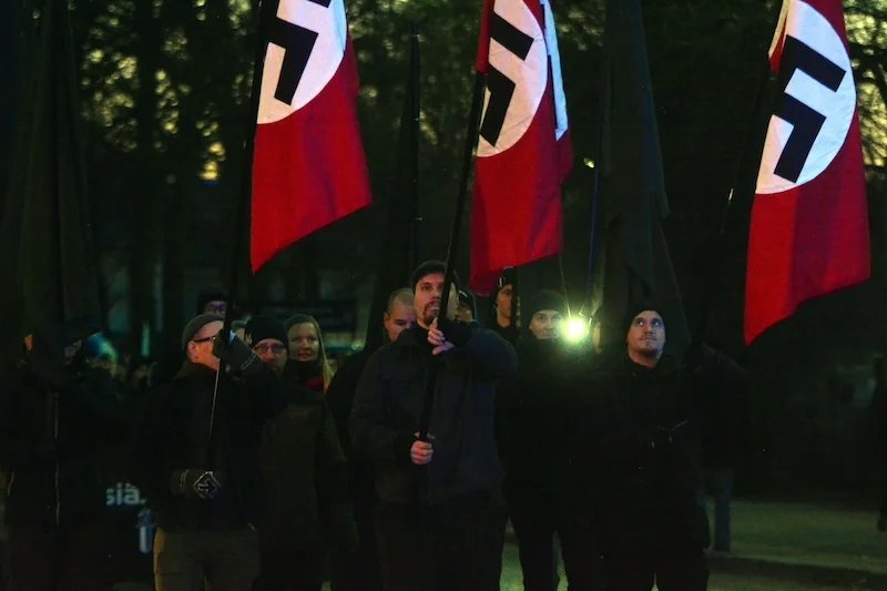 Men marching in Nazi uniforms and carrying Nazi flags.