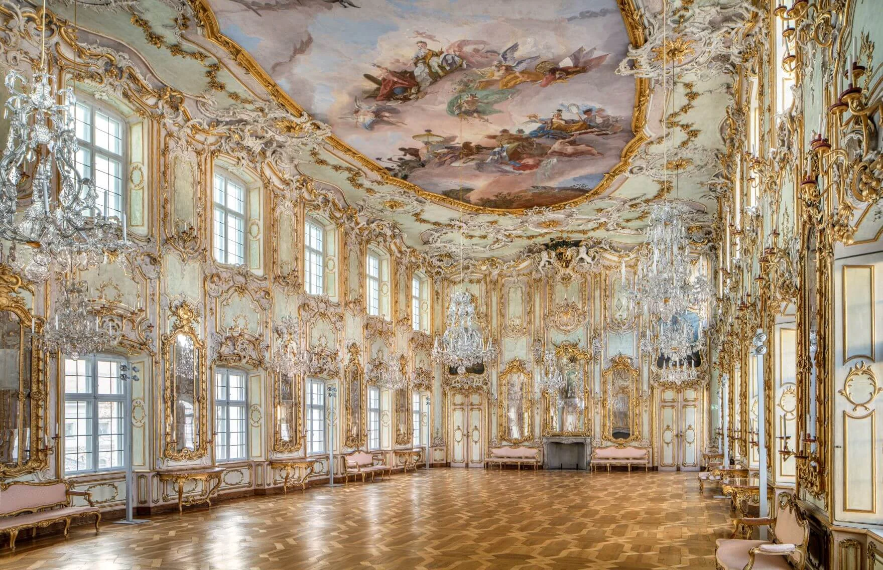 An ornate ballroom with gilded decorations, large windows, chandeliers, a parquet floor, and a painted ceiling depicting a classical scene.