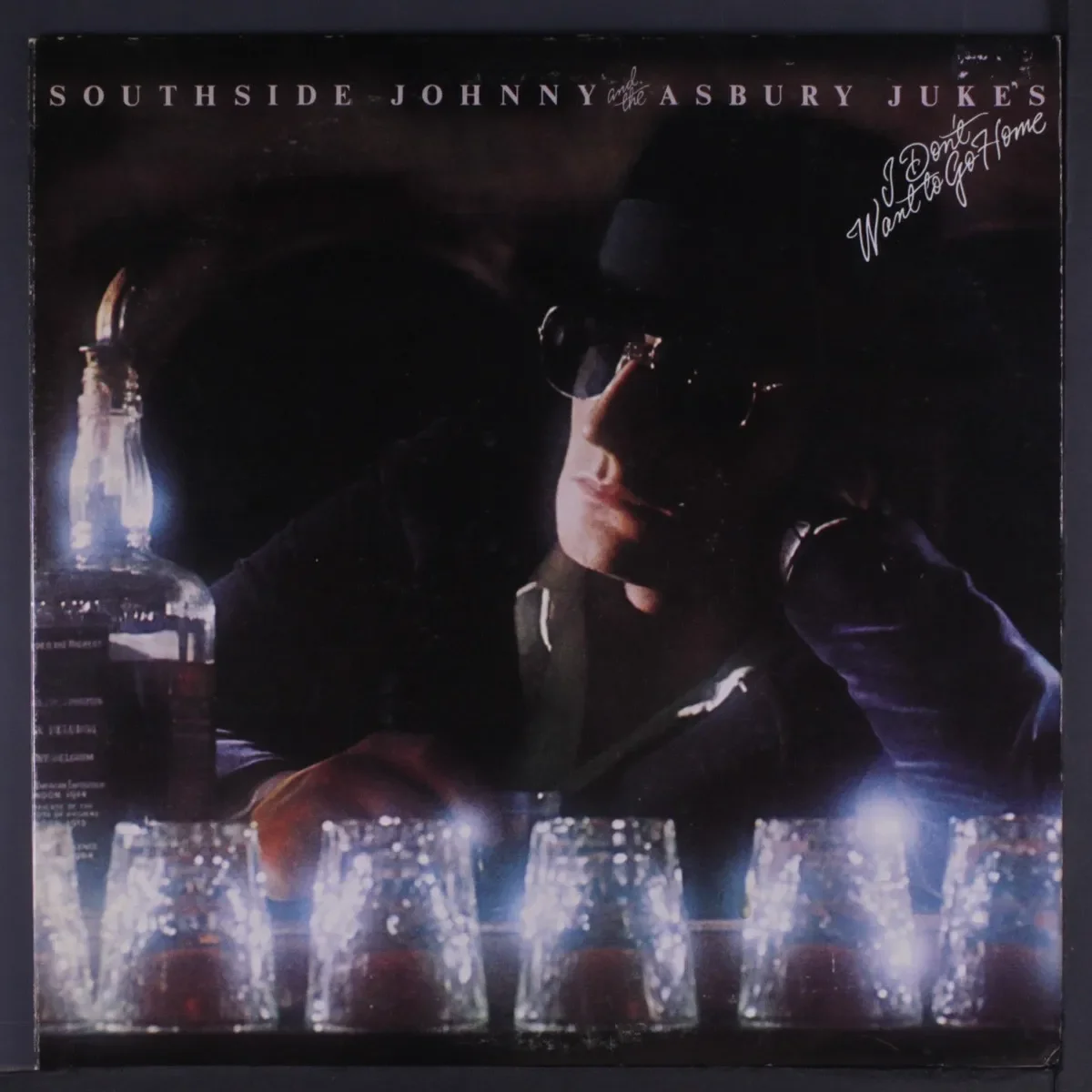 Southside Johnny in sunglasses sitting at a dimly lit bar with a bottle and glasses, conveying a reflective mood.