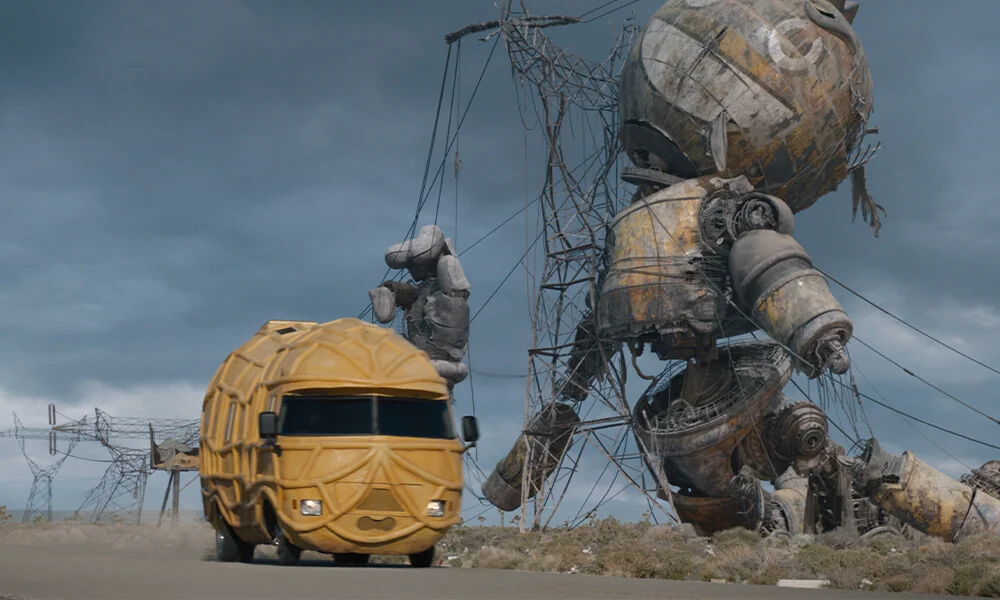 A peanut-shaped vehicle drives on a road under a cloudy sky, passing a giant, rusted robot slumped against tangled metal structures, creating a surreal scene.