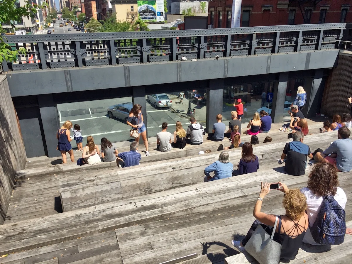 People sit on wooden bleachers viewing a street below through large windows. It’s sunny, creating a relaxed, open-air urban atmosphere.