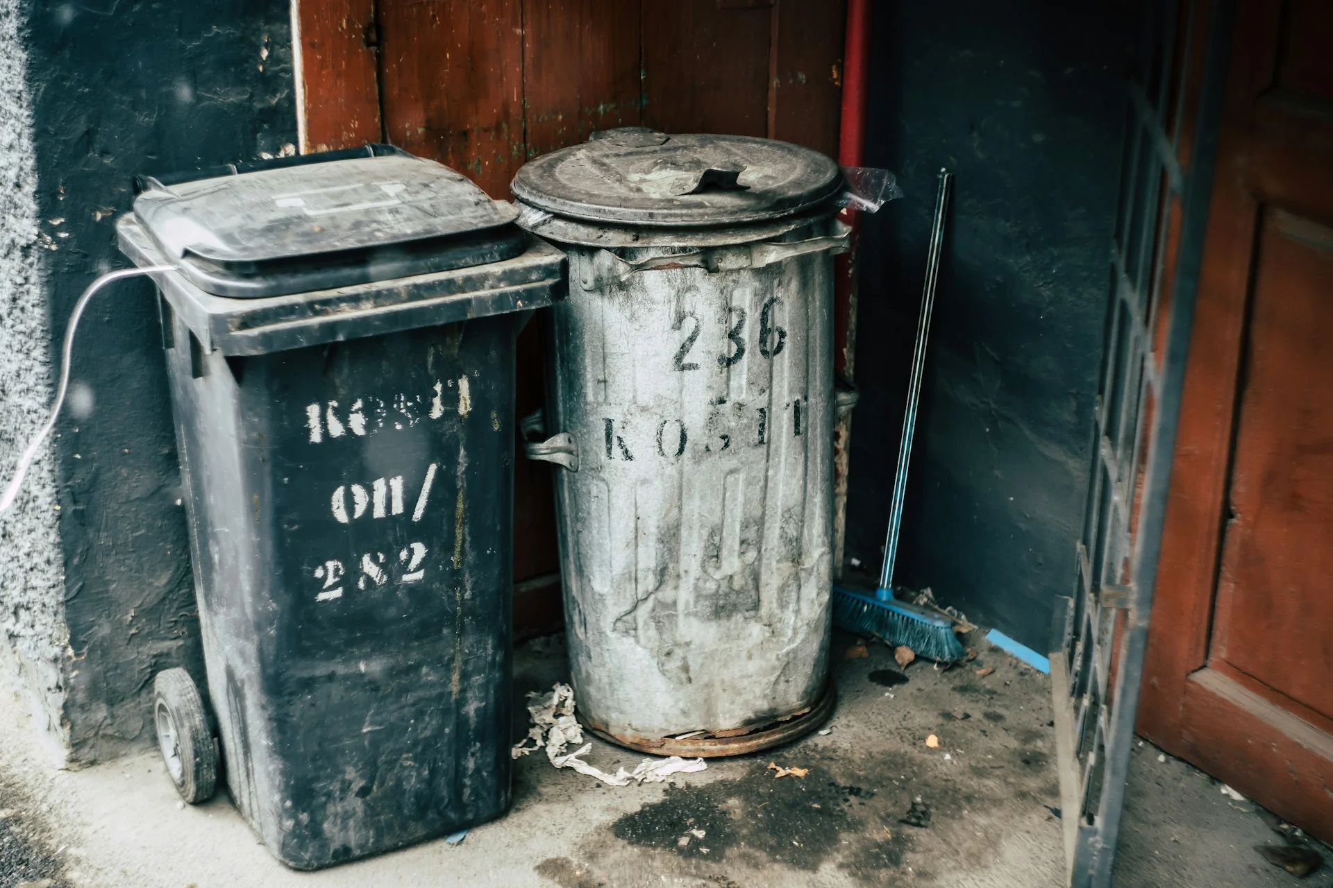 Two trash bins, one black with a lid and one metallic, stand against a weathered wall. A broom leans beside them, suggesting a gritty urban setting.