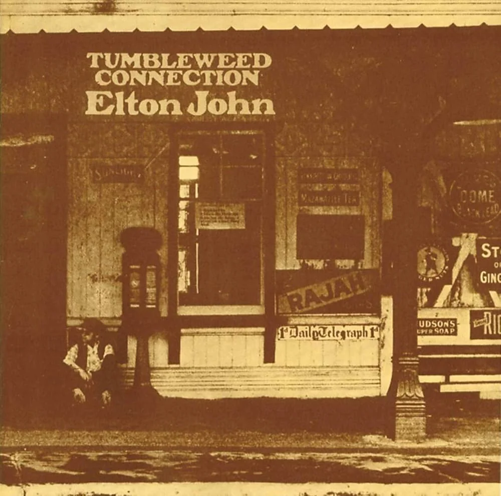 Album cover. Sepia-toned image of a vintage storefront with old-fashioned signs. A person sits on the left, creating a nostalgic, rustic atmosphere.