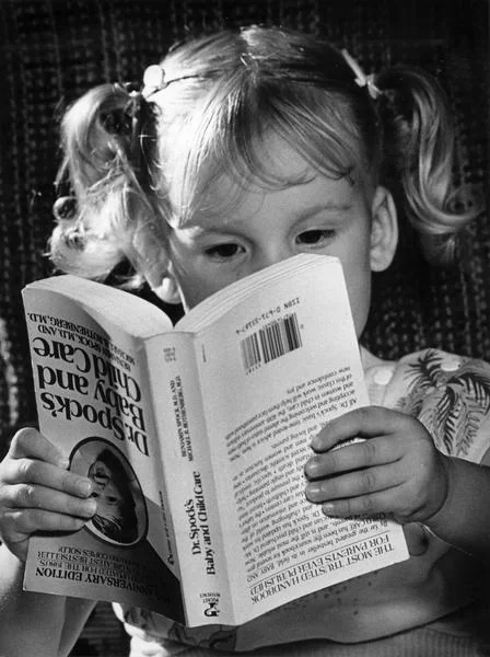 Black and white photo of a little girl reading Dr. Spock's Baby and Childcare. She is holding the book upside down."